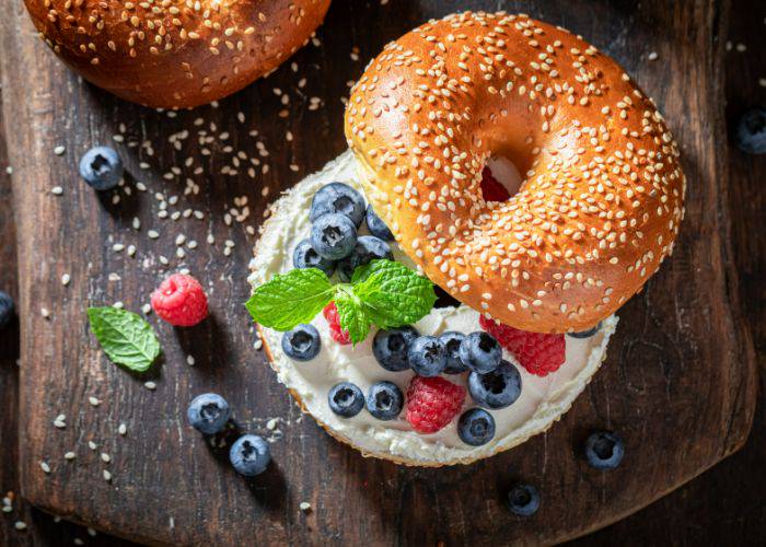 A top-down shot of a bagel on a dark wood board. The top is slightly askew, revealing cream, blueberries, raspberries and mint.